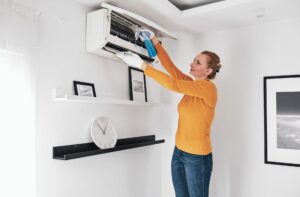 Woman cleaning wall-mounted air conditioner unit in modern living room, emphasizing HVAC maintenance and home comfort in Woods Cross.